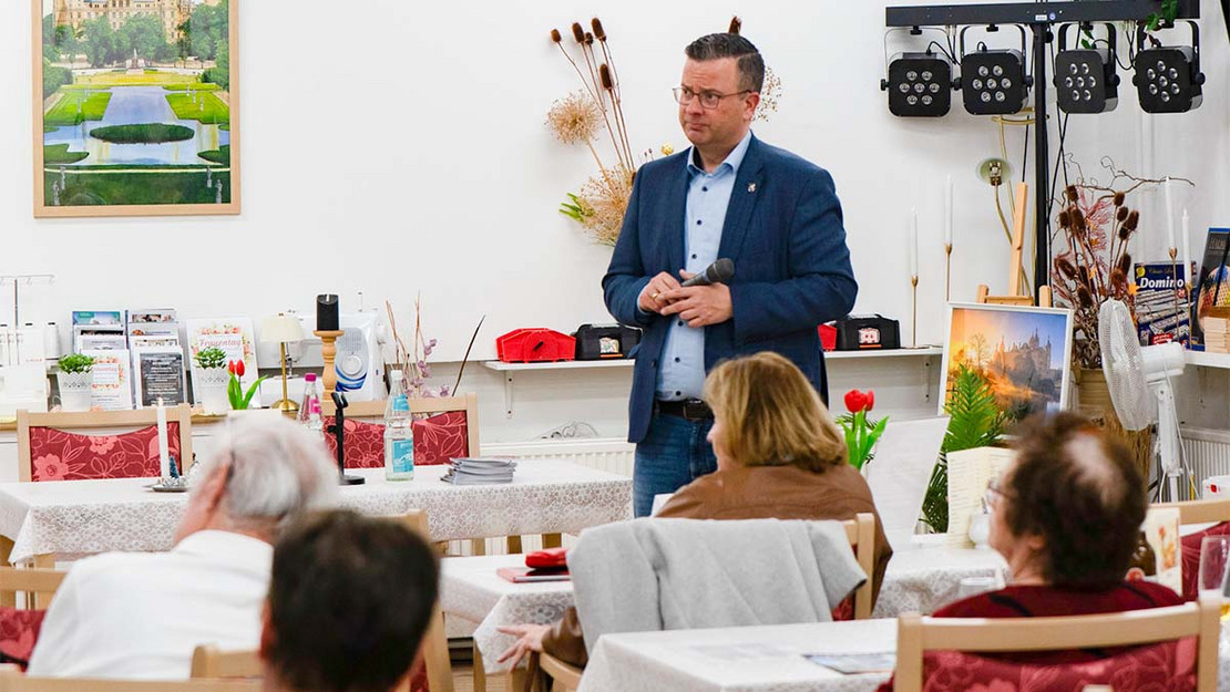 Ein Mann steht in einem Veranstaltungssaal, mehrere Personen sitzen auf Stühlen und hören zu. 
