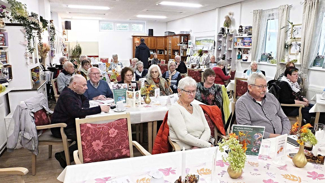 Mehrere Menschen sitzen an Tischen und hören einem Vortrag zu. 