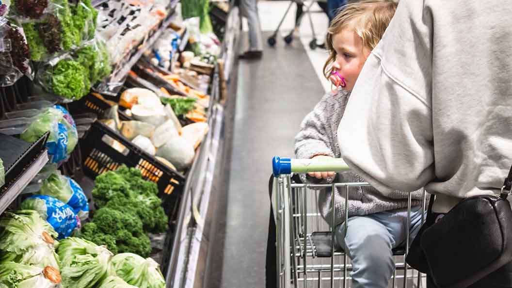 Kind im Einkaufswagen vor dem Gemüseregal im Supermarkt.